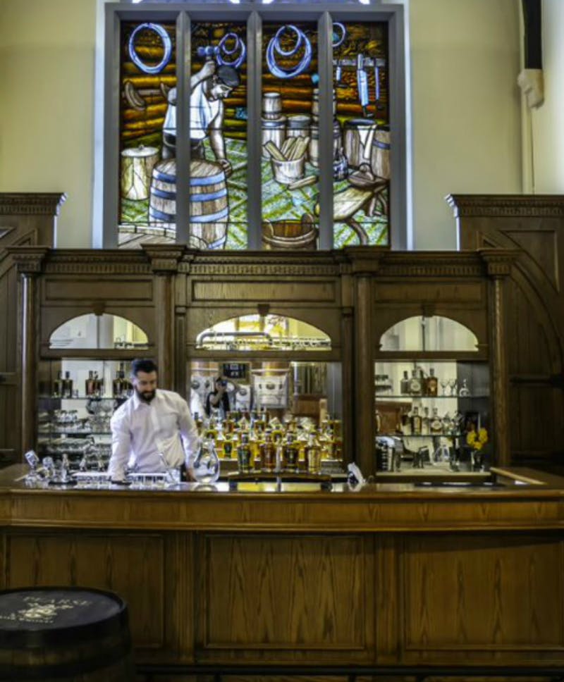 ワイン樽とブドウを描いたステンドグラスの窓の下に、様々なボトルとグラスを備えた木製のバーの後ろにバーテンダーが立っています。