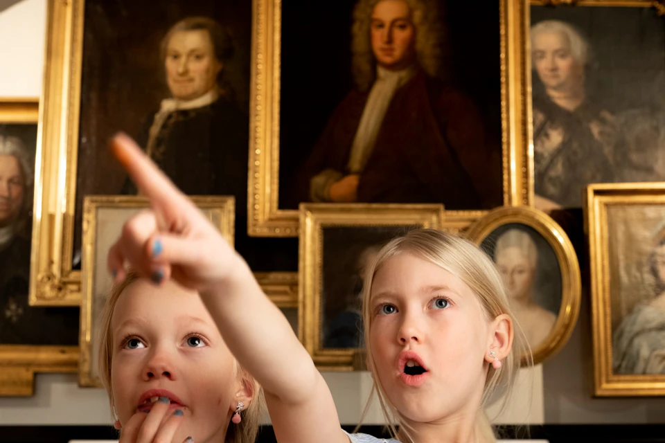 Two young girls in a museum, one pointing and the other looking surprised, stand in front of several framed portraits.
