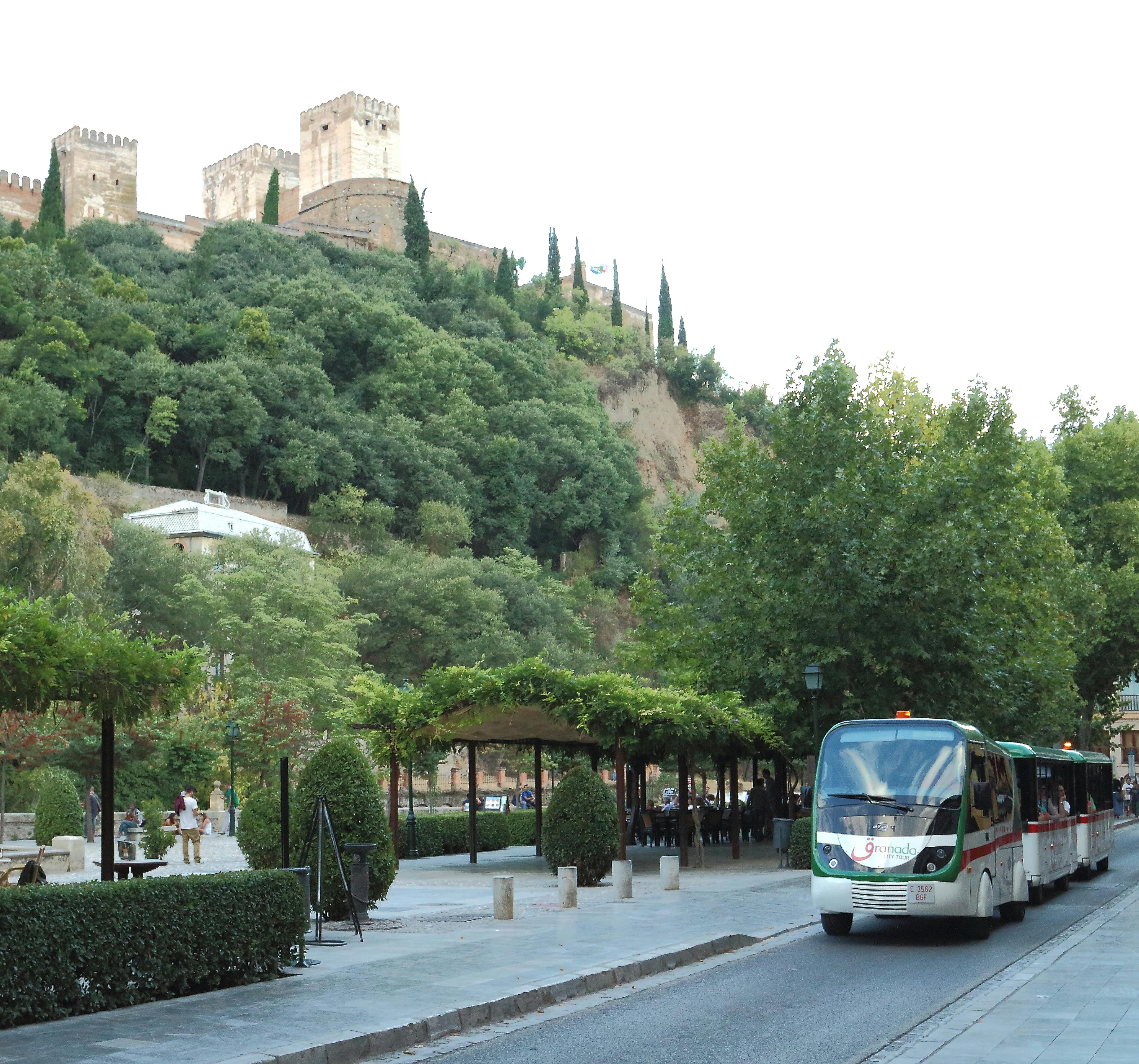 울창한 녹음과 나무를 지나면 길거리 수준의 관광 트램이 운행되며, 언덕 꼭대기에는 유서 깊은 성이 배경으로 보입니다.
