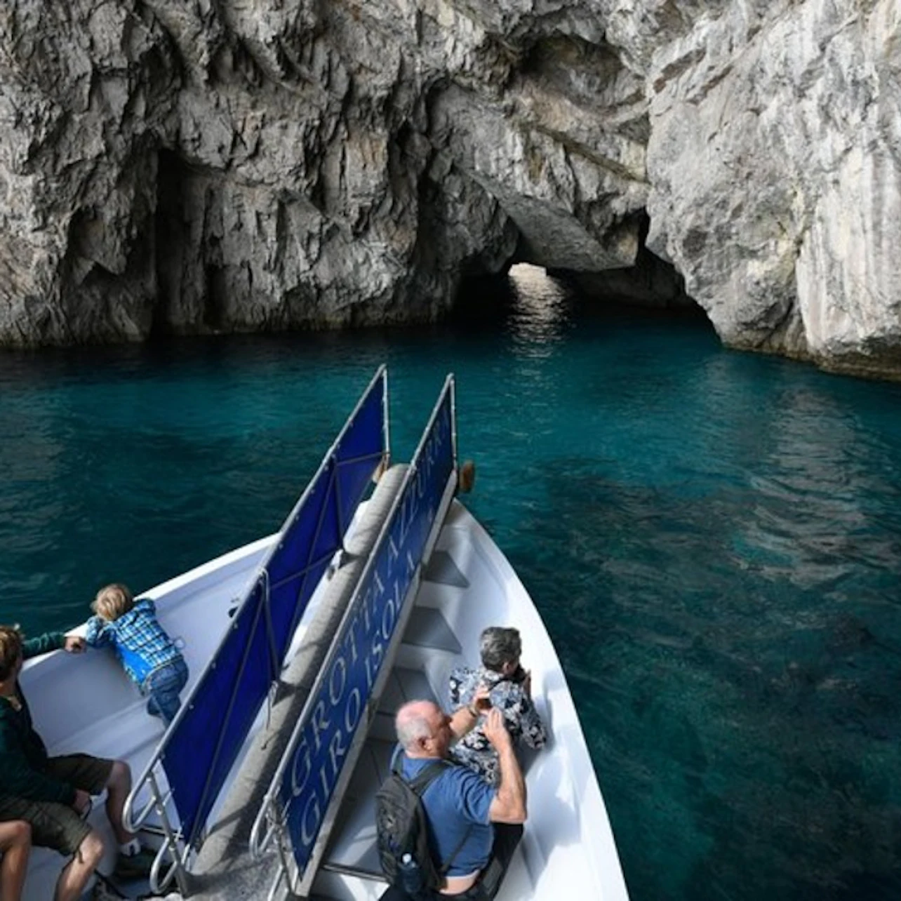 People on a boat approach a rocky cave entrance over clear blue water. Some are taking photos while one child leans over the railing.