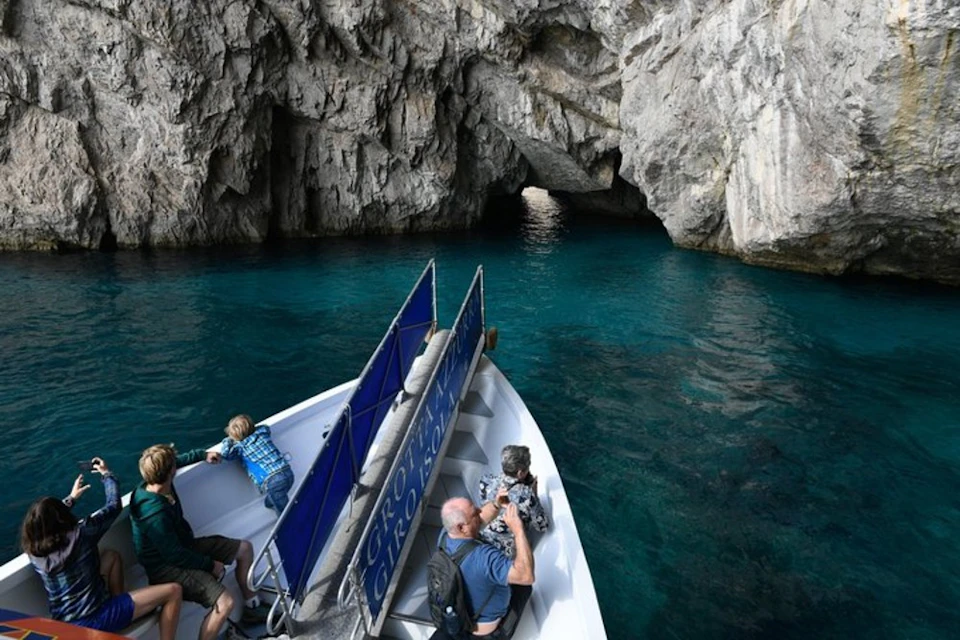 People on a boat approach a rocky cave entrance over clear blue water. Some are taking photos while one child leans over the railing.