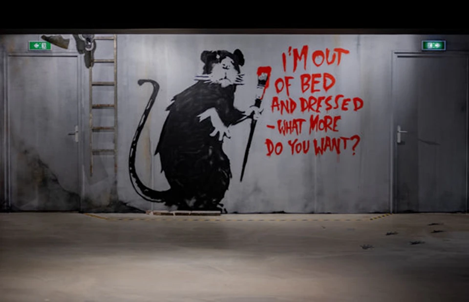 Graffiti of a large rat holding a paintbrush with red paint, next to text reading "I'M OUT OF BED AND DRESSED -WHAT MORE DO YOU WANT?".