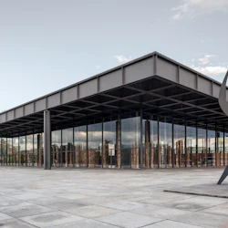 Modern byggnad med stora glasfönster, platt överhängande tak och en abstrakt metallskulptur på ett rymligt kaklat torg.