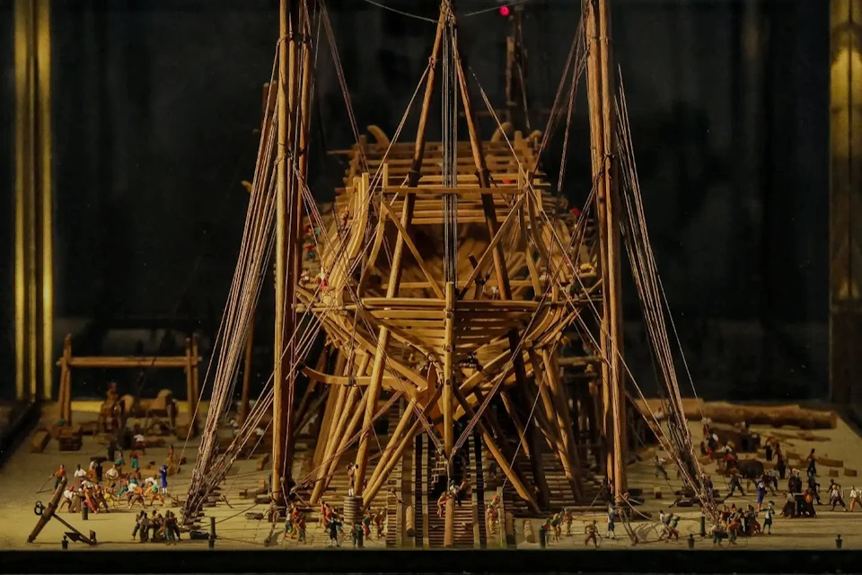 A detailed wooden ship under construction with miniature figures working around it, surrounded by scaffolding and rigging.