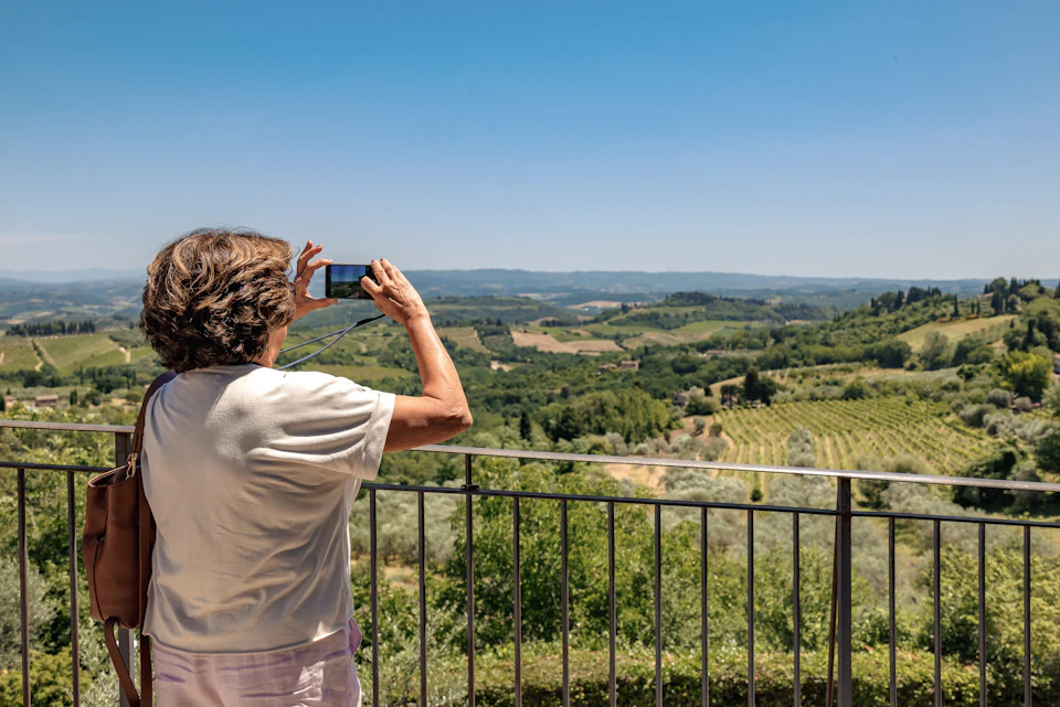 A person taking a photo of a scenic landscape featuring hills, fields, and trees from a balcony under a clear blue sky.