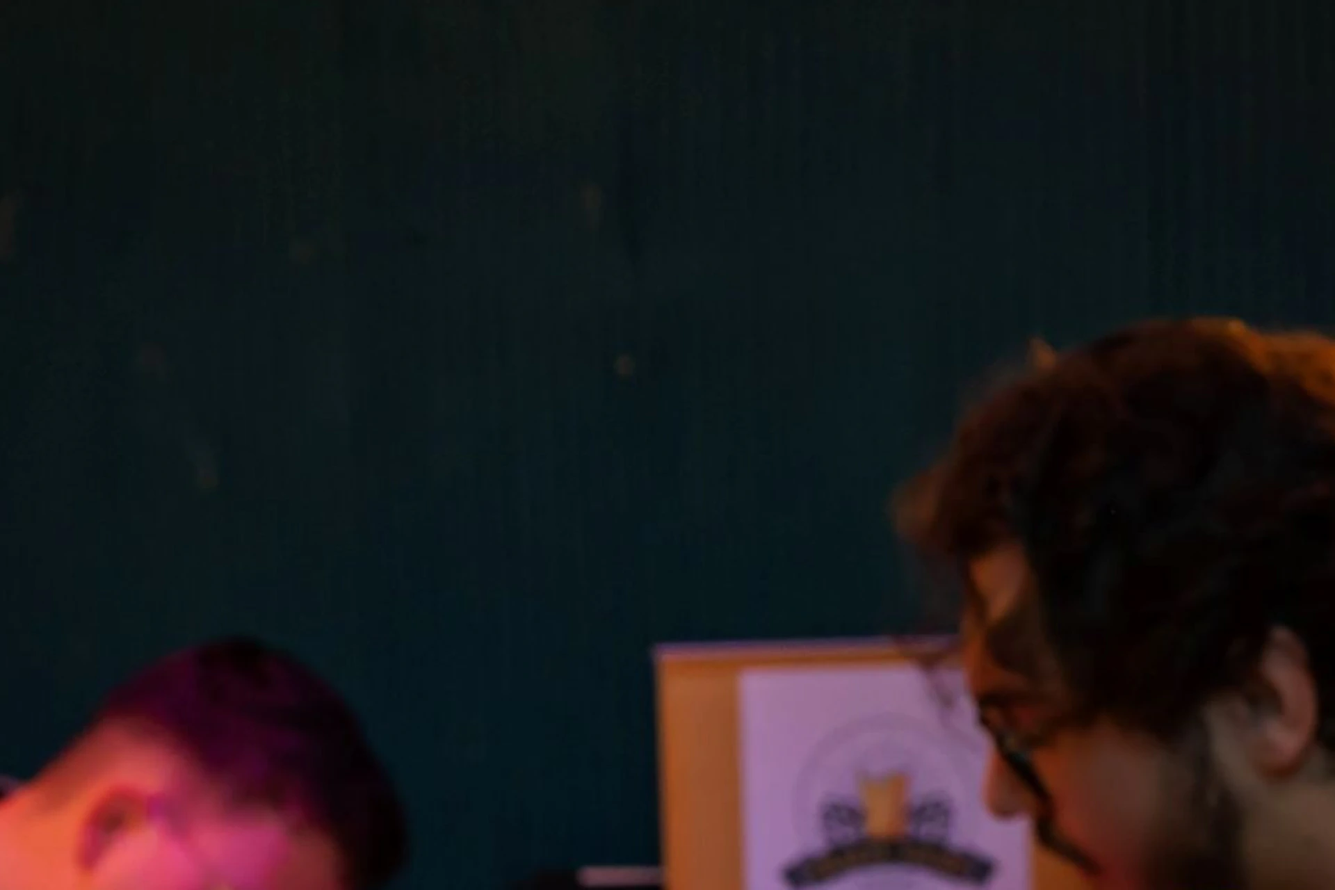 A man in a yellow shirt holding a glass in a dimly lit room with people sitting at a table in the background.