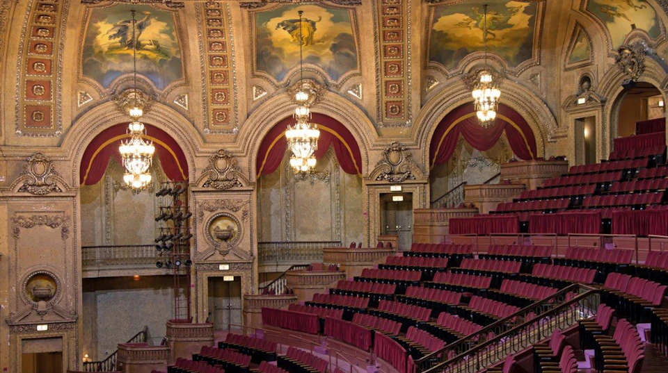 Ornate theater interior with red velvet seats, chandeliers, decorative arches, and painted ceiling murals.