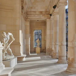 Utsmyckad hall med stenpelare och två klassiska statyer i välvda alkover, en till vänster och en i bakgrunden.