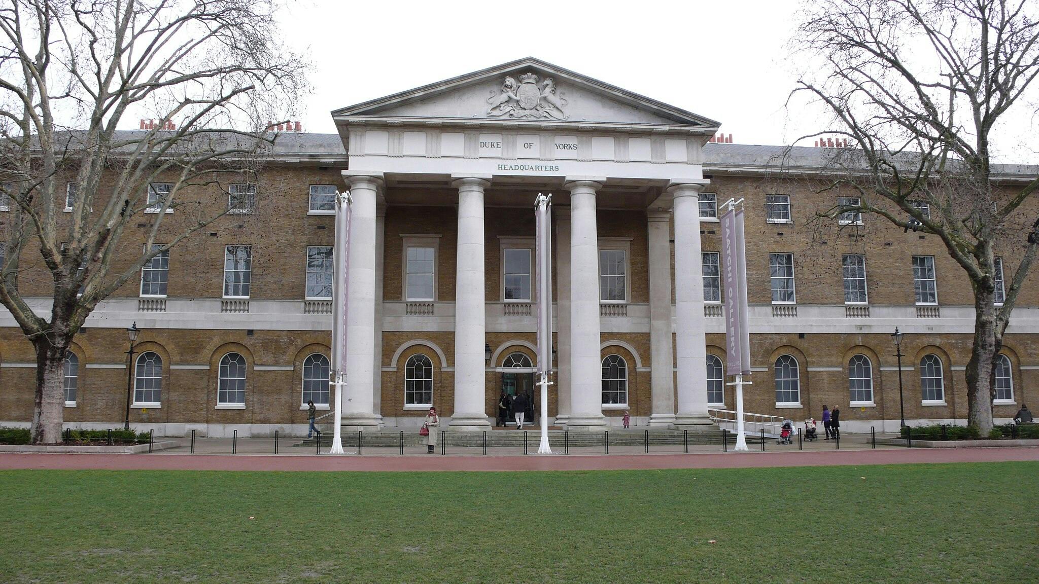 Un grande edificio con la scritta "DUKE OF YORKS HEADQUARTERS", colonne, finestre ad arco, alberi spogli e persone vicino all'ingresso.