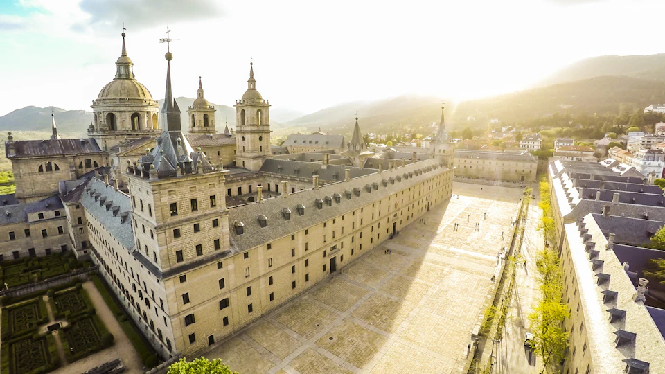 Historic stone building complex with towers, situated in a spacious square under a bright, hazy sky. Mountains in the background.