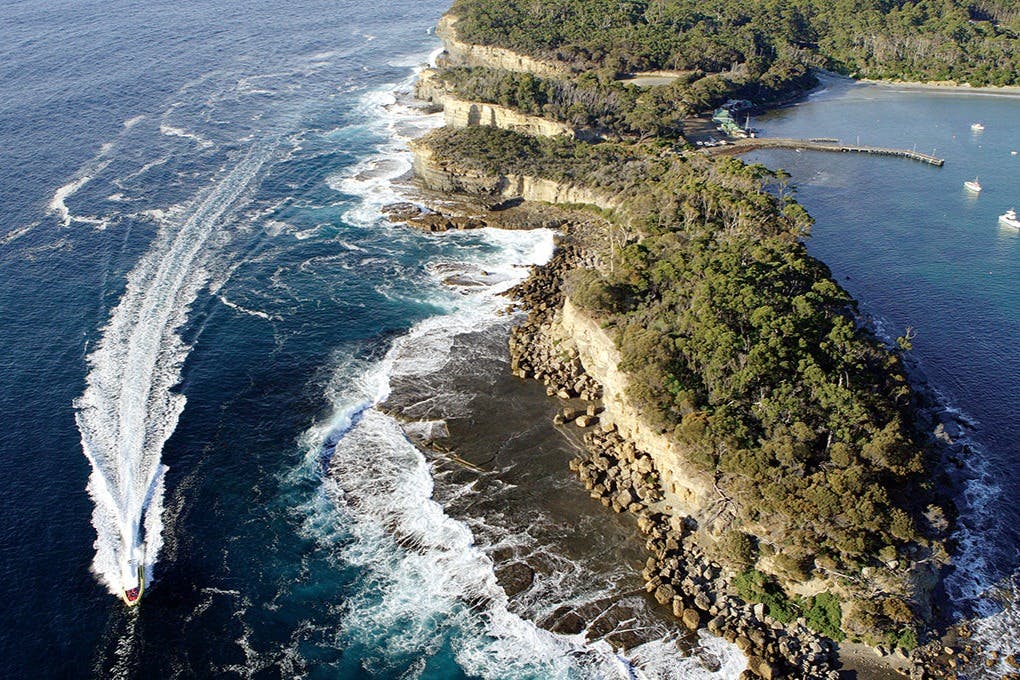 Letecký pohled na členité pobřeží se skalnatými útesy, rozbouřenými vlnami a bujnou zelenou vegetací pod jasnou oblohou.