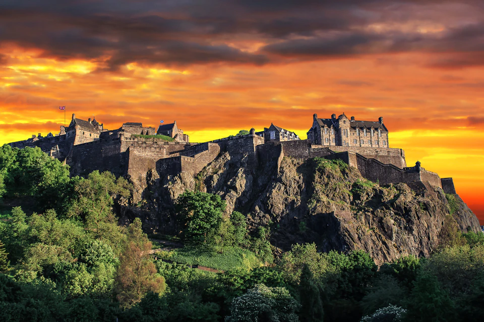 A historic castle sits atop a rocky hill, surrounded by lush greenery, under a vibrant orange and yellow sunset sky.