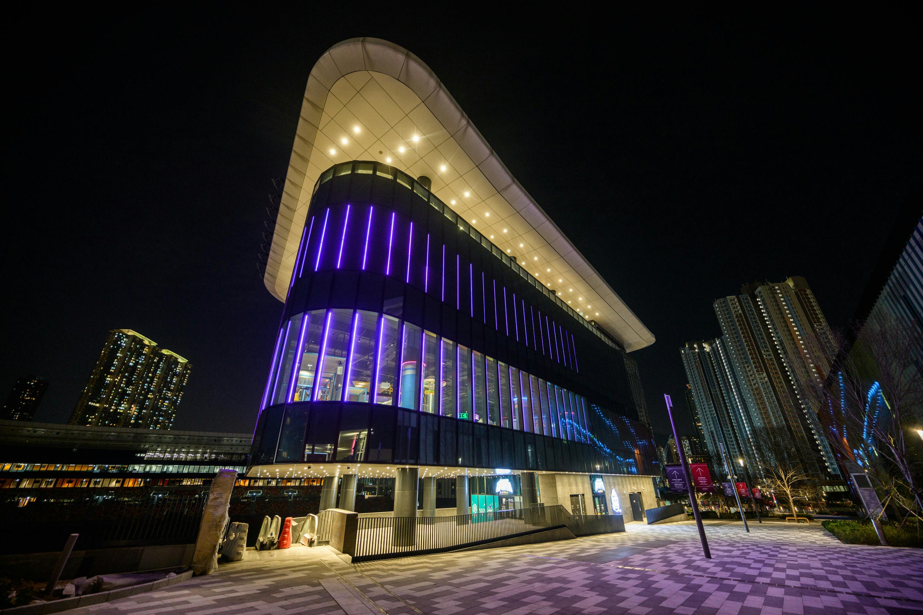 A modern, illuminated building with colorful vertical lights and a curved roof, set against a nighttime cityscape.