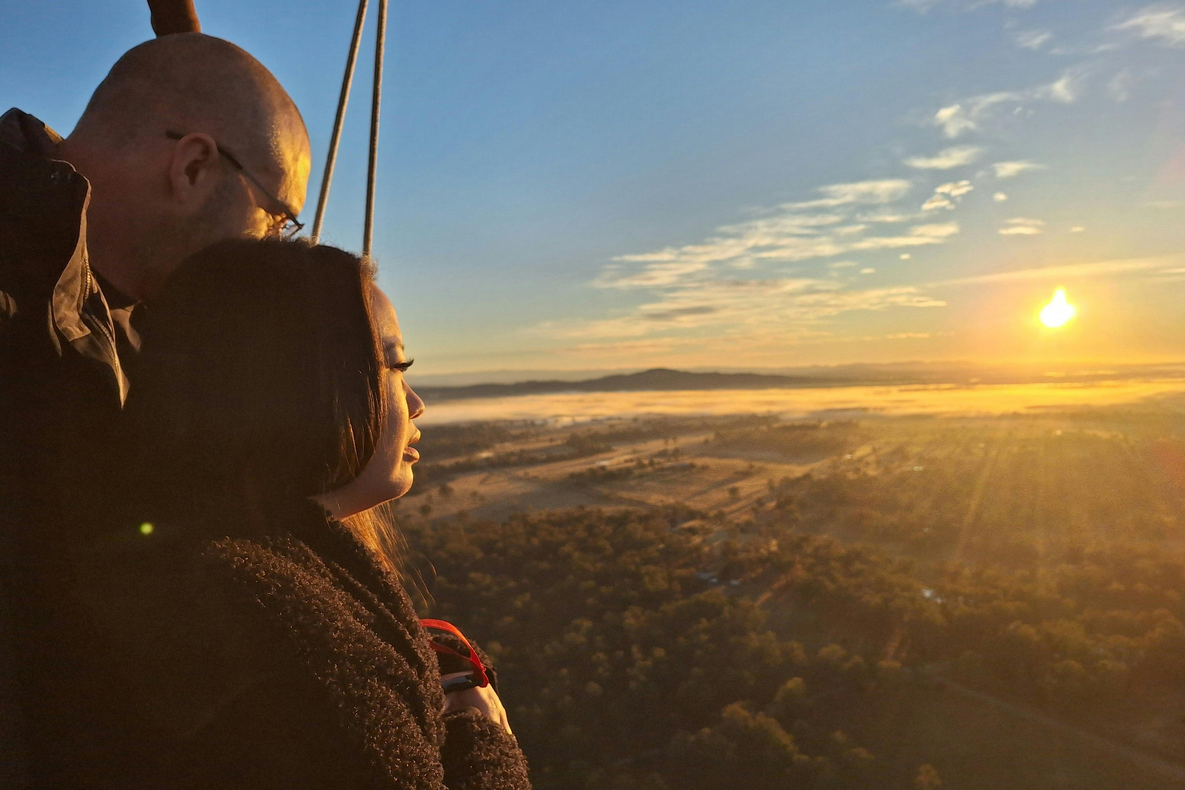 Zwei Personen in einem Heißluftballonkorb genießen den Blick auf den Sonnenaufgang über einer Landschaft mit Feldern, Bäumen und fernen Hügeln.