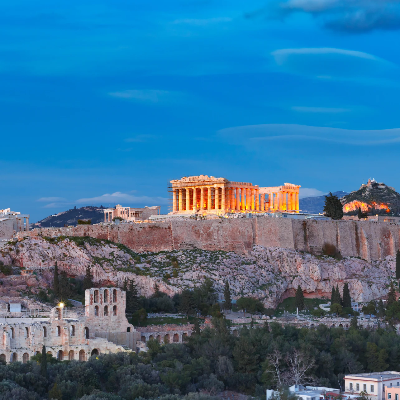 The illuminated Parthenon atop the Acropolis in Athens, Greece, viewed from a distance against a twilight sky.