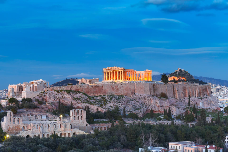 The illuminated Parthenon atop the Acropolis in Athens, Greece, viewed from a distance against a twilight sky.