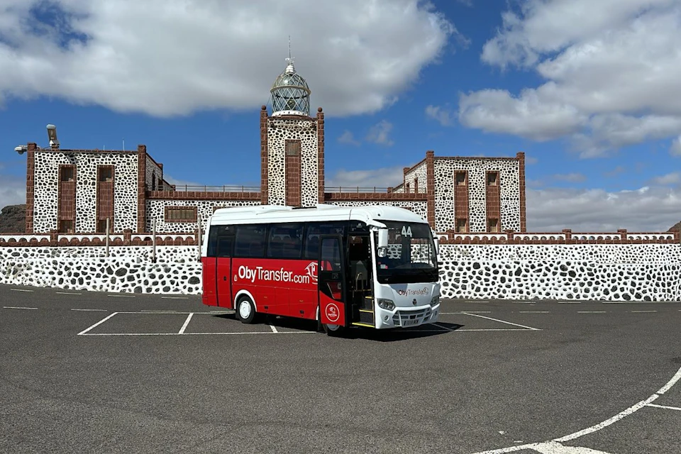 Red and white tour bus parked in front of a stone building with a tower and clock, under a partly cloudy sky.