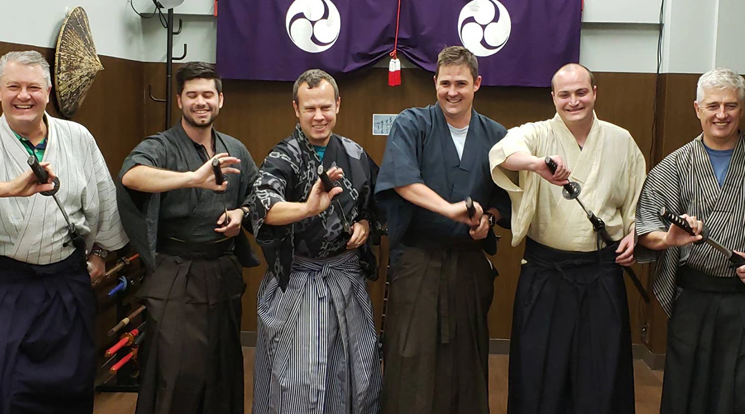 Three people wearing traditional Japanese attire and holding small objects in a room with purple banners and wooden decor.