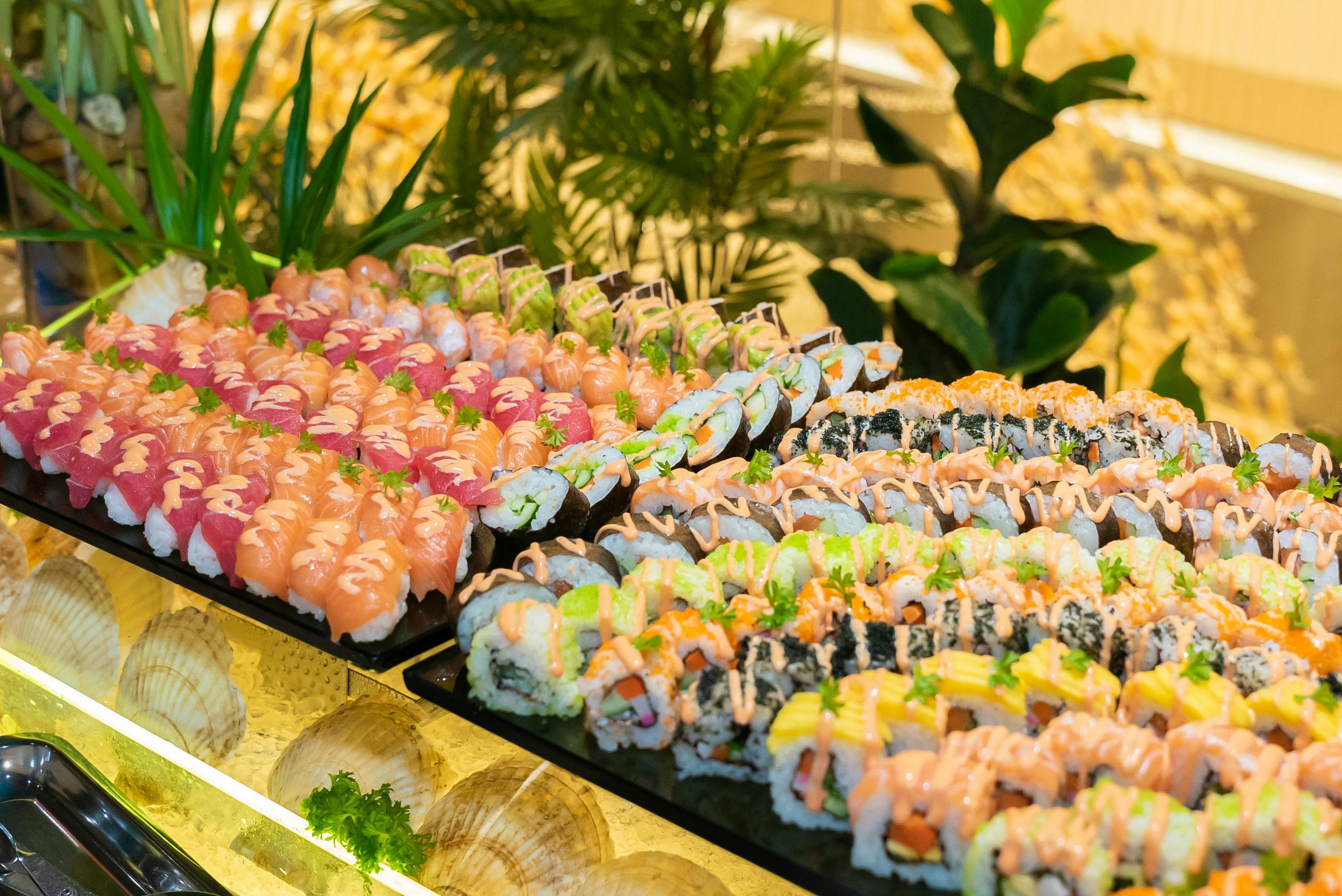 An arrangement of colorful sushi rolls displayed on trays with garnishes, set against a decorative background with plants.