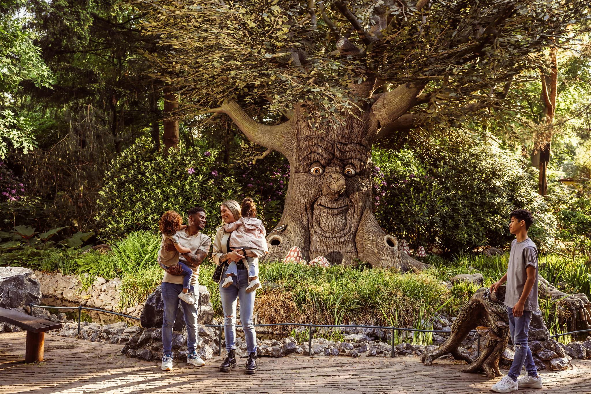 Eine Familie mit zwei Kindern steht in einer parkähnlichen Umgebung vor einem großen Baum, in den ein Gesicht geschnitzt ist.