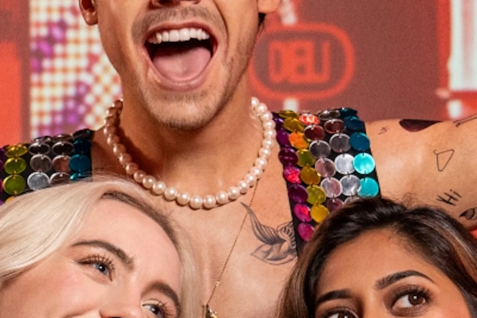 Two women smile and pose for a selfie with a wax statue of a man in a colorful outfit in a vibrant, illuminated setting.