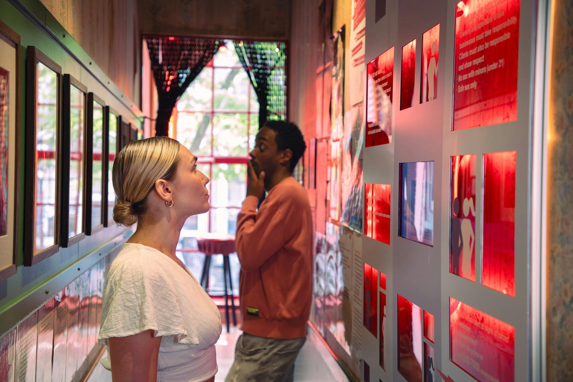 Due persone osservano opere d'arte incorniciate e testi a parete in uno spazio stretto e ben illuminato della galleria con una grande finestra in fondo.