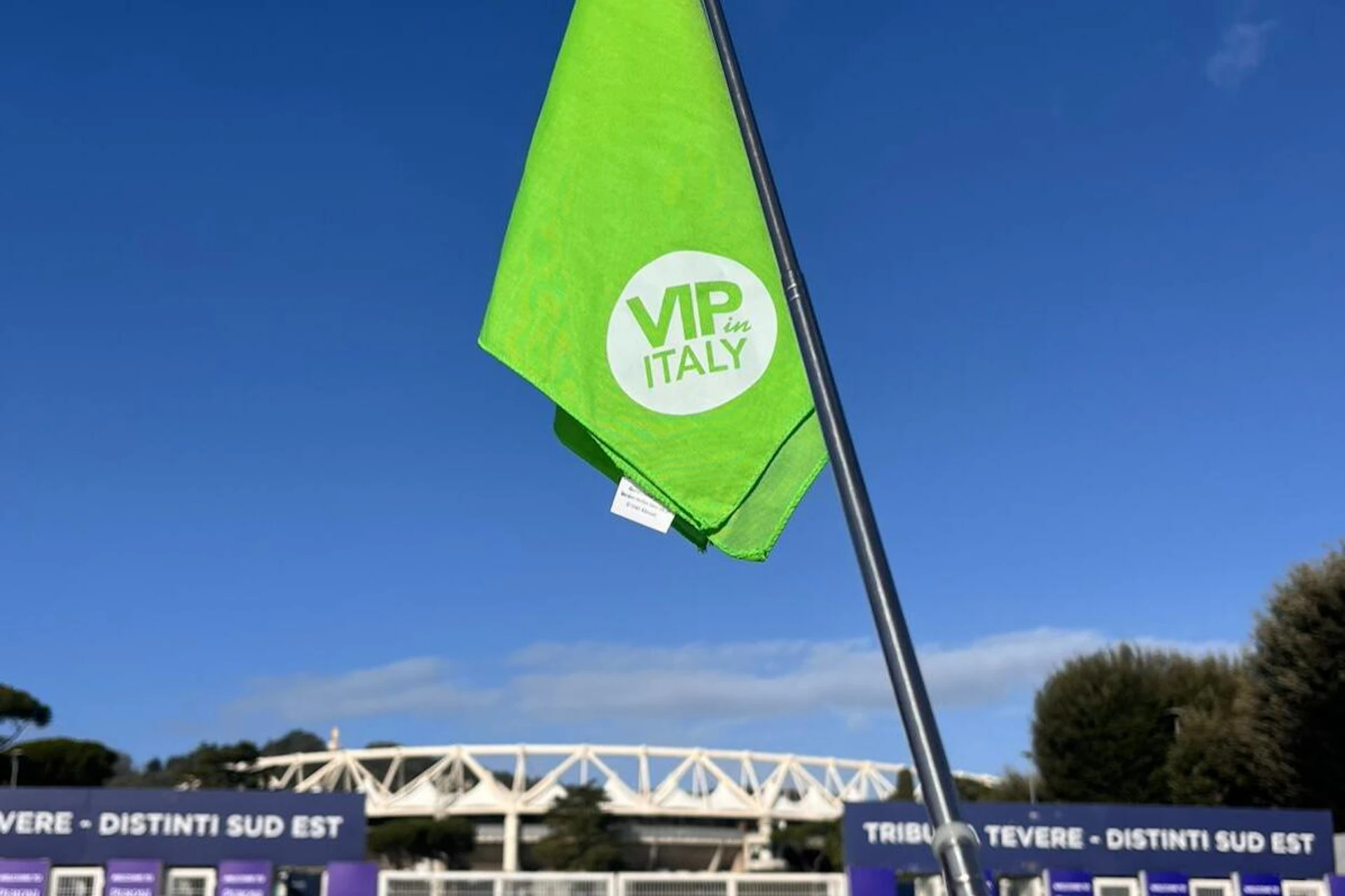 A green flag with the text 'VIP in Italy' on it, mounted on a pole, with a stadium and blue sky in the background.