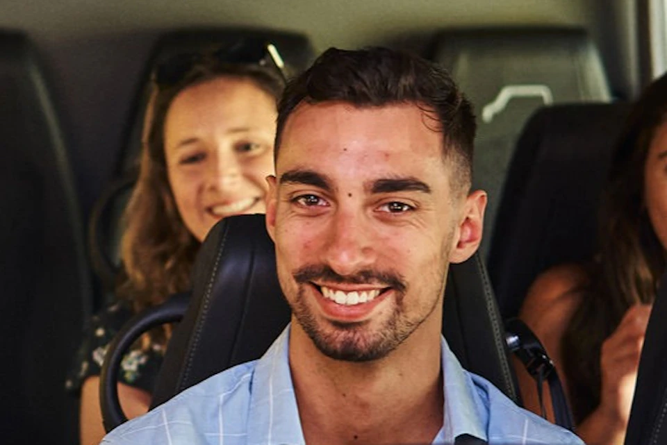 A group of people smiling while seated inside a vehicle, with visible rows of seats and a window to the right.