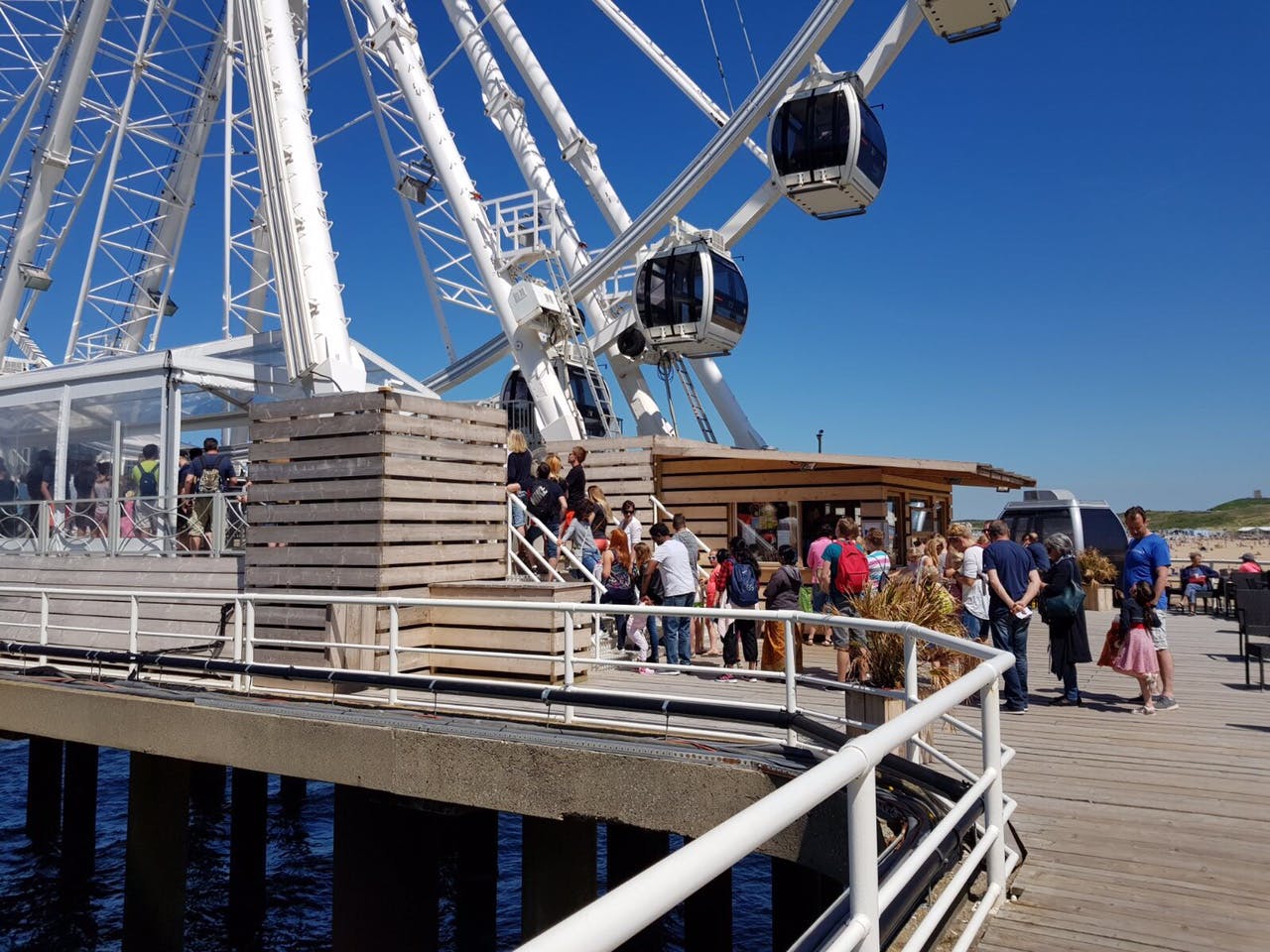 Lidé čekající ve frontě u paty velkého ruského kola poblíž dřevěné budovy za slunečného dne u vody.