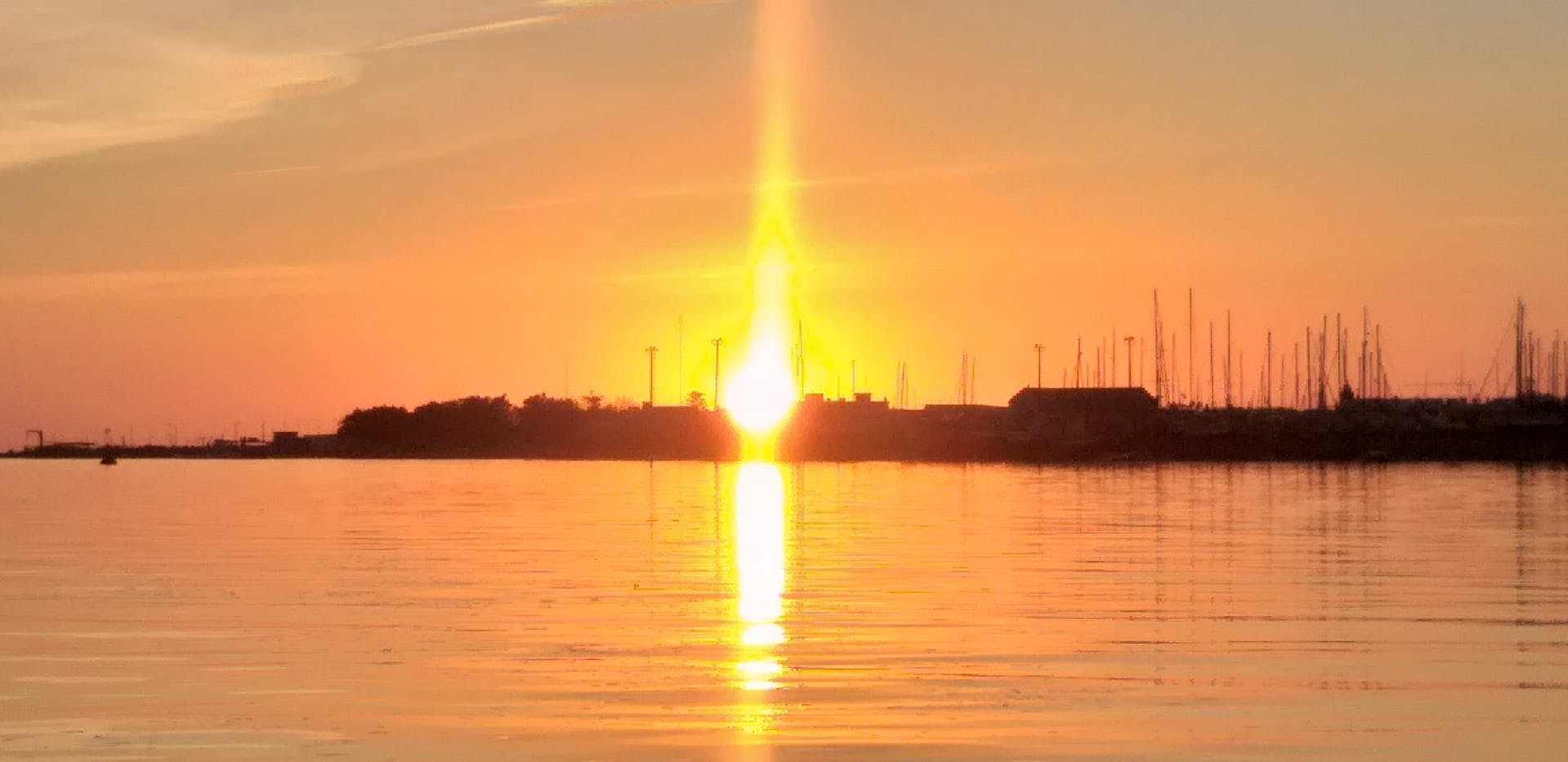 Солнце садится над спокойным водоемом, излучая золотистое сияние и отражая на поверхности силуэты зданий и столбов.