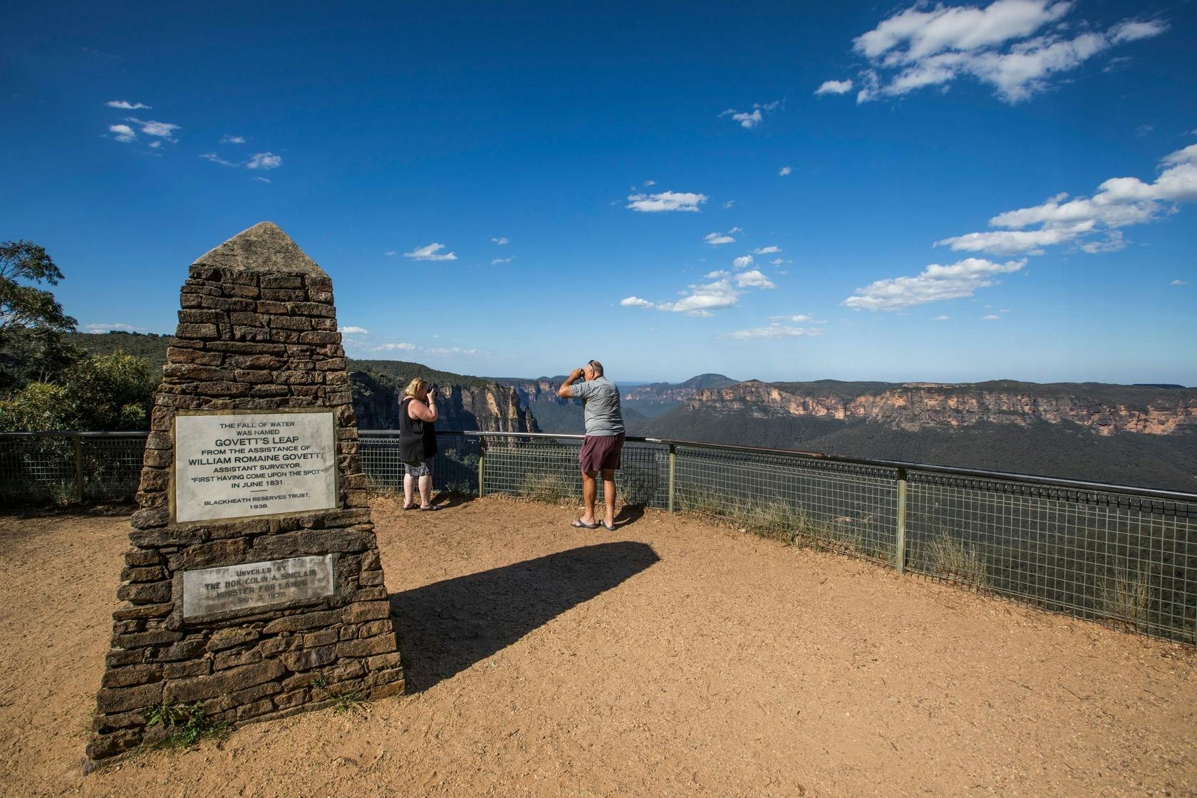 Two people at a scenic lookout with a plaque and a railing, overlooking a mountain range under a clear sky.