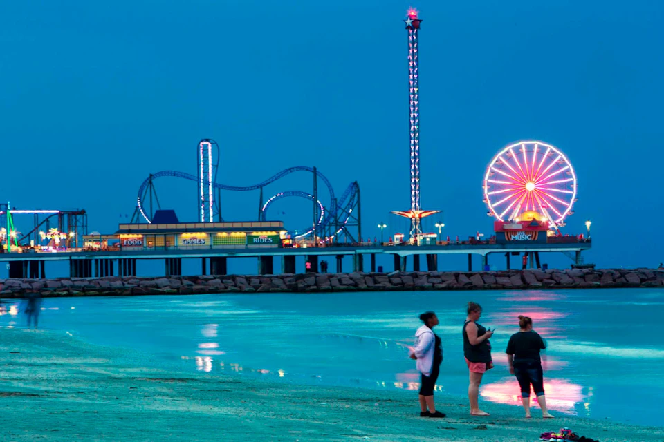 A beach scene at dusk with three people standing near the water, and an amusement park with rides visible in the background.
