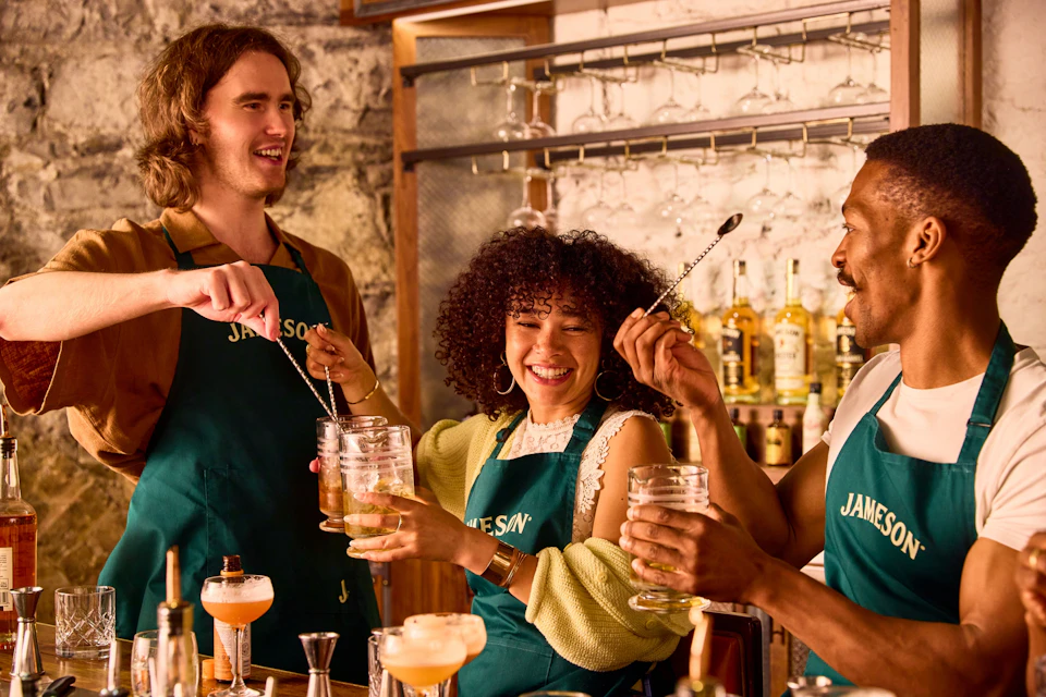 Two people wearing green aprons smiling and preparing drinks at a bar, surrounded by cocktail glasses and tools.