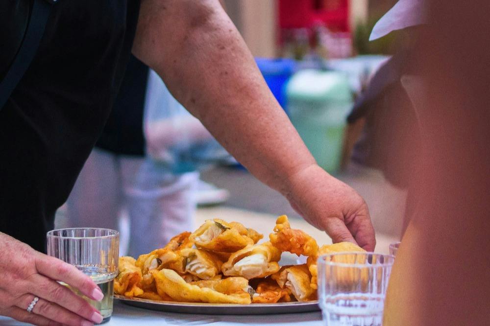 A person reaches for a food item on a plate of fried pastries, with glasses of liquid nearby on the table.