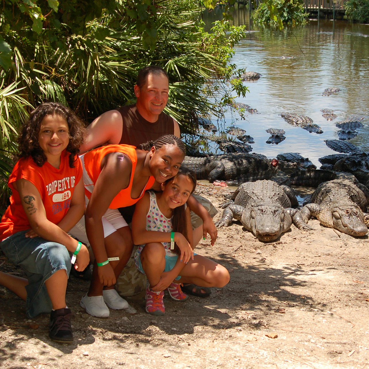 Four people posing near a pond with numerous alligators; lush vegetation and water in the background.