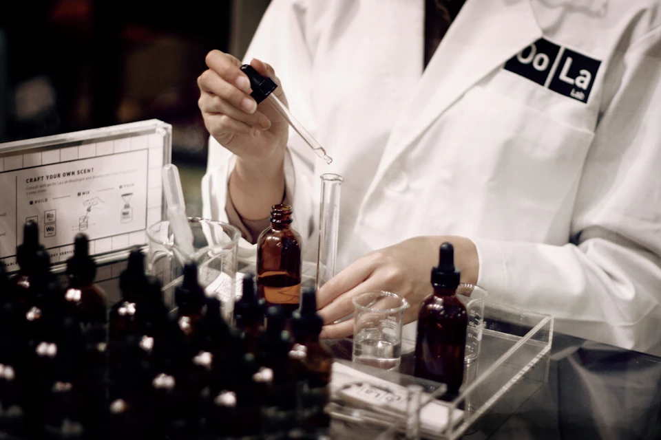Person in a lab coat using a dropper to transfer liquid into a test tube, surrounded by amber bottles and lab equipment.