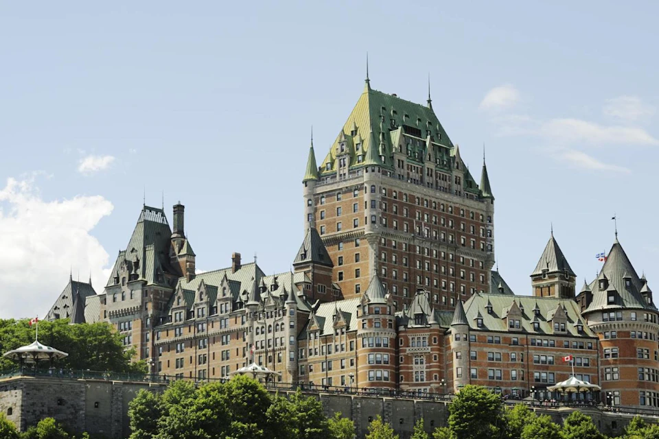 Large historic building with green roofs and spires, surrounded by trees, under a clear blue sky.