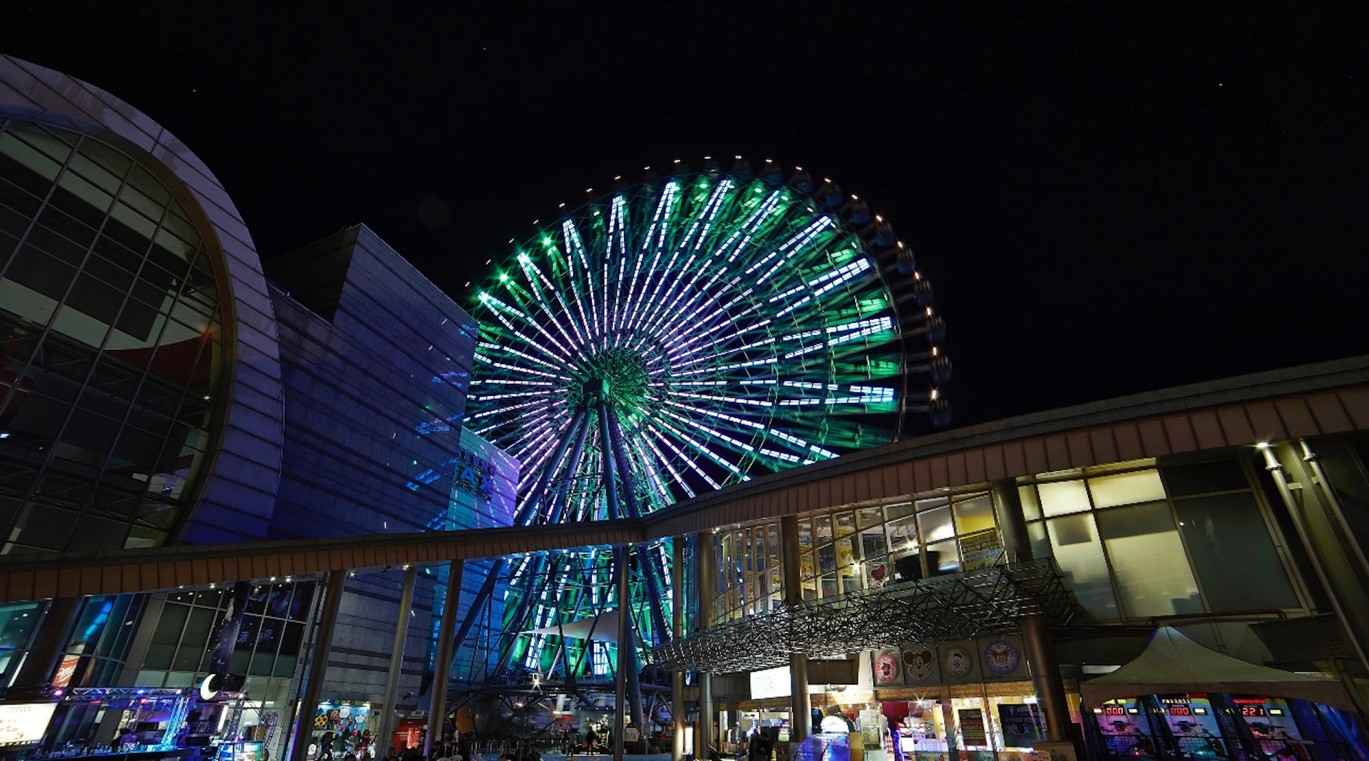 An illuminated Ferris wheel with green and purple lights at night, with nearby buildings and shops in the foreground.