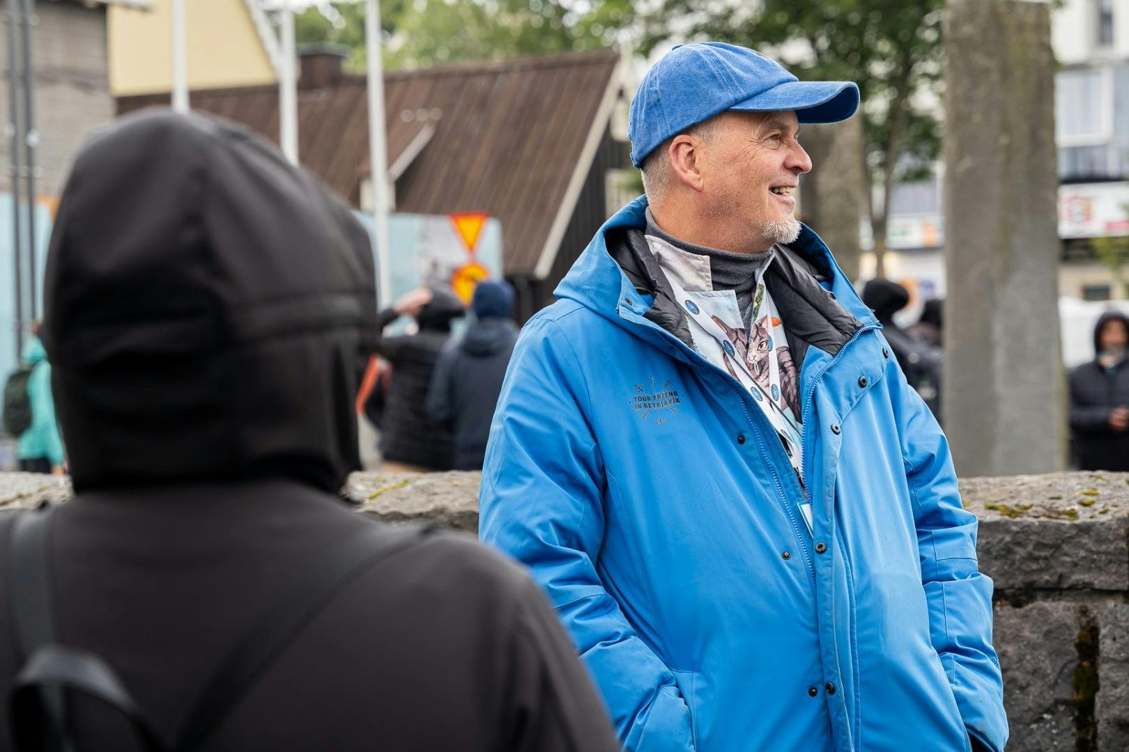 파란색 재킷과 모자를 쓴 남성이 돌담 근처 야외에 서 있으며, 배경에는 사람과 건물이 보입니다.