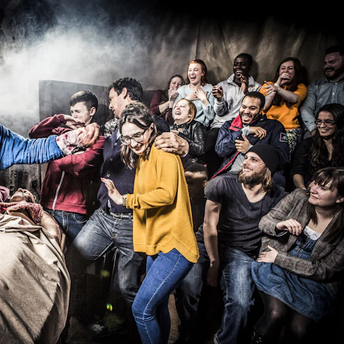 En grupp människor reagerar med nöje och chock när en utklädd artist som håller falska organ uppträder i en mörk, teatralisk miljö med rök.