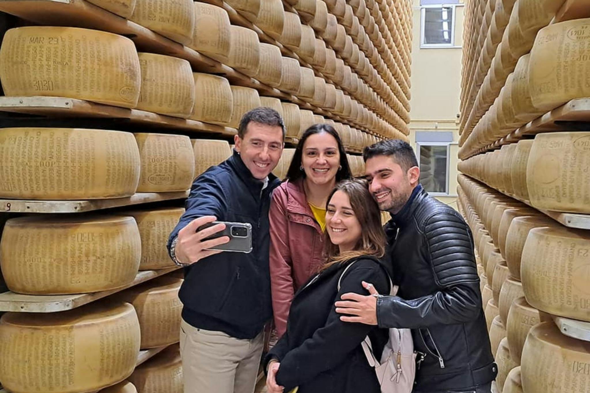Quattro persone, sorridenti, si scattano un selfie in una stanza piena di scaffali impilati con grandi forme di formaggio.