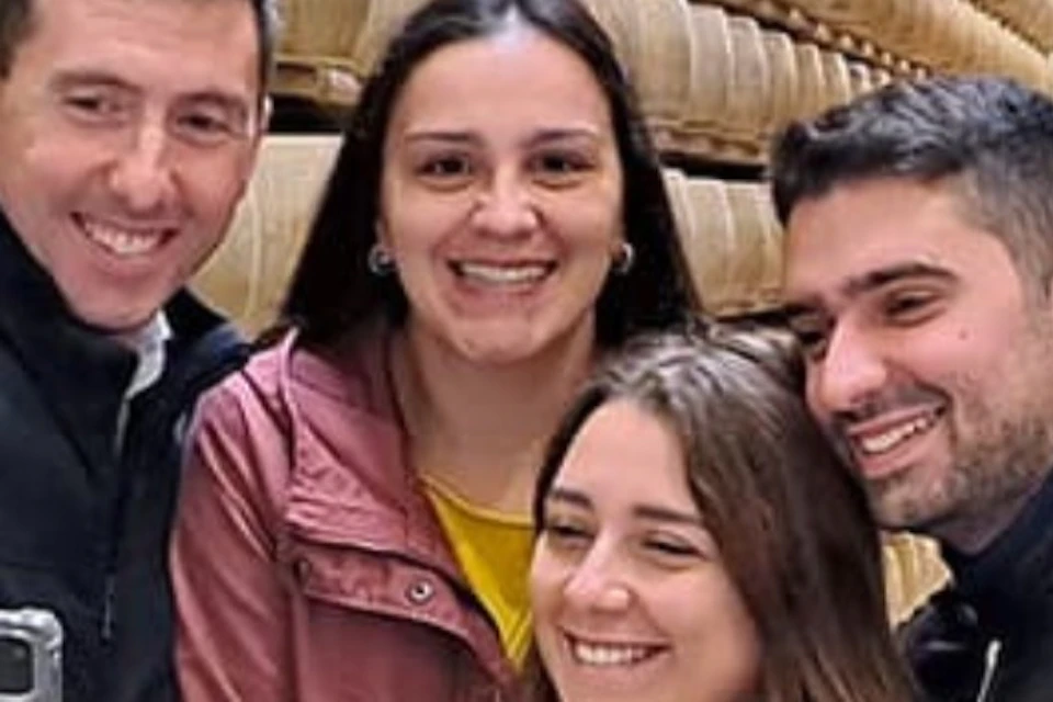 Four people, smiling, taking a selfie in a room filled with shelves stacked with large wheels of cheese.