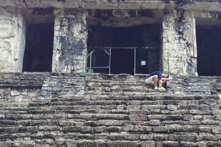 A person crouching on stone steps in front of an ancient stone structure with a dark interior.