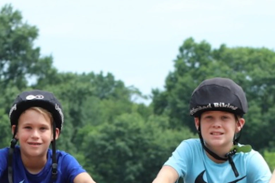 Five people wearing helmets, standing with bicycles on a bridge with trees and ornate stone railings in the background.
