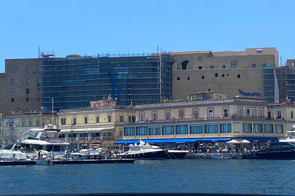 Boats docked in a marina in front of buildings, some under construction, with scaffolding and a clear, blue sky above.