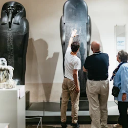 Three people examine an ancient Egyptian sarcophagus displayed in a museum, with inscriptions and carvings visible.