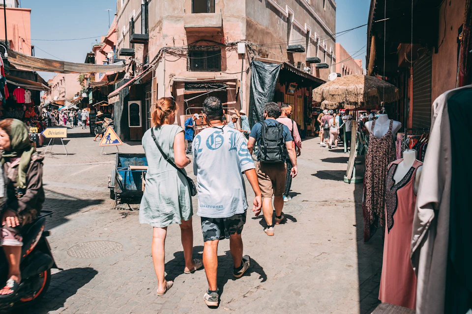 People walking through a busy market street with rustic buildings, shops, and various goods on display under a sunny sky.