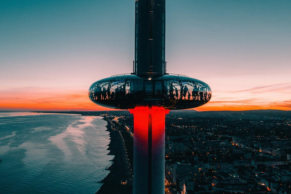 A futuristic observation tower with a reflective capsule illuminated by red light, overlooking a coastline at sunset.