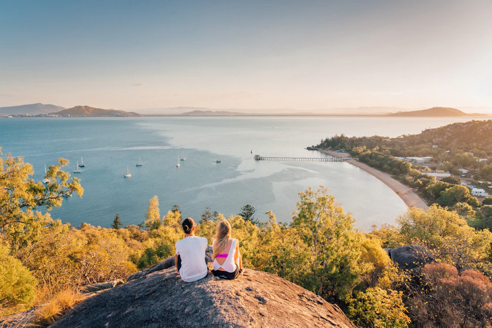 Two people sit on a rock overlooking a scenic bay with boats, a pier, and surrounding greenery under a clear sky.