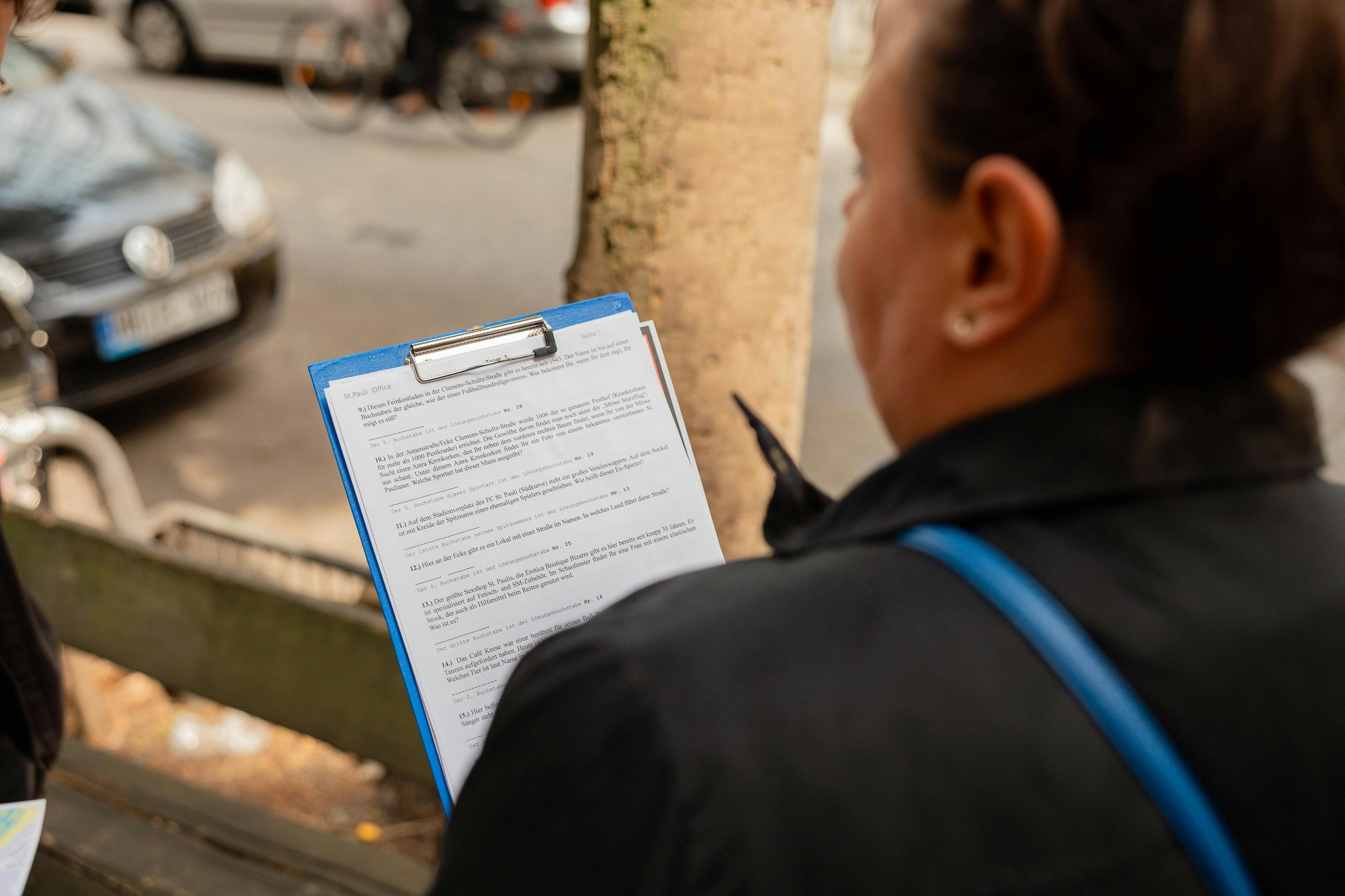 一个人拿着一张带纸的蓝色剪贴板，在街上阅读，街上停着汽车，背景模糊，还有一辆自行车。